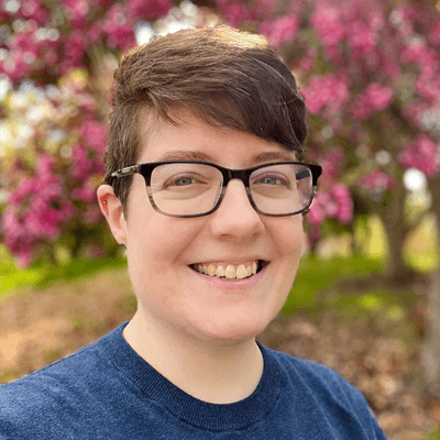 Elh poses in front of a blooming purple tree outside on a sunny day. They wear a blue shirt, dark rimmed glasses, and short hair.