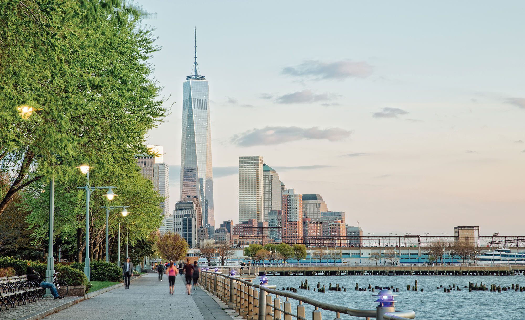 People walking and running on a sidewalk next to a river in New York City with One World Trade Center in the background.