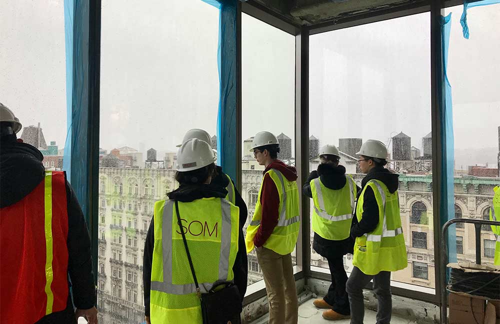 A group of students wearing hardhats and high visibility jackets inside of an under-construction building overlooking a cityscape.