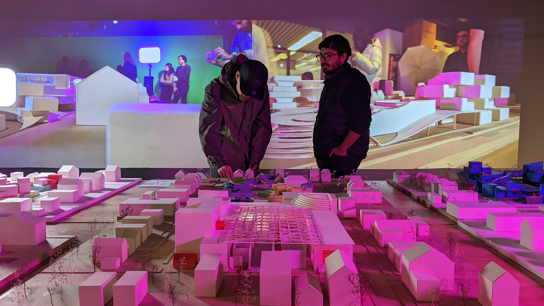Two people look at a small model neighborhood on display in the TVLab. A digital display is lit up behind them.