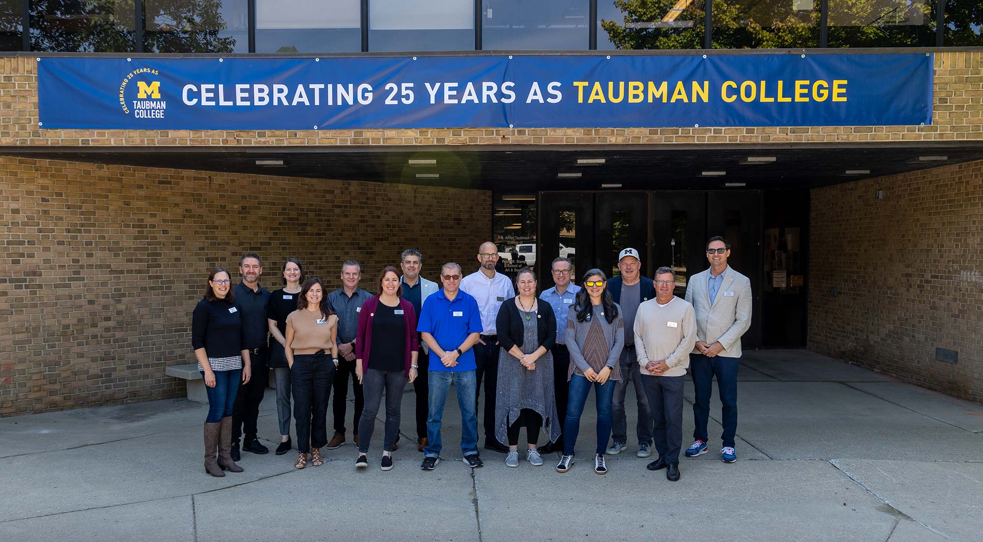 A group of people standing in front of the Art + Architecture Building with a banner celebrating 25 years of Taubman College