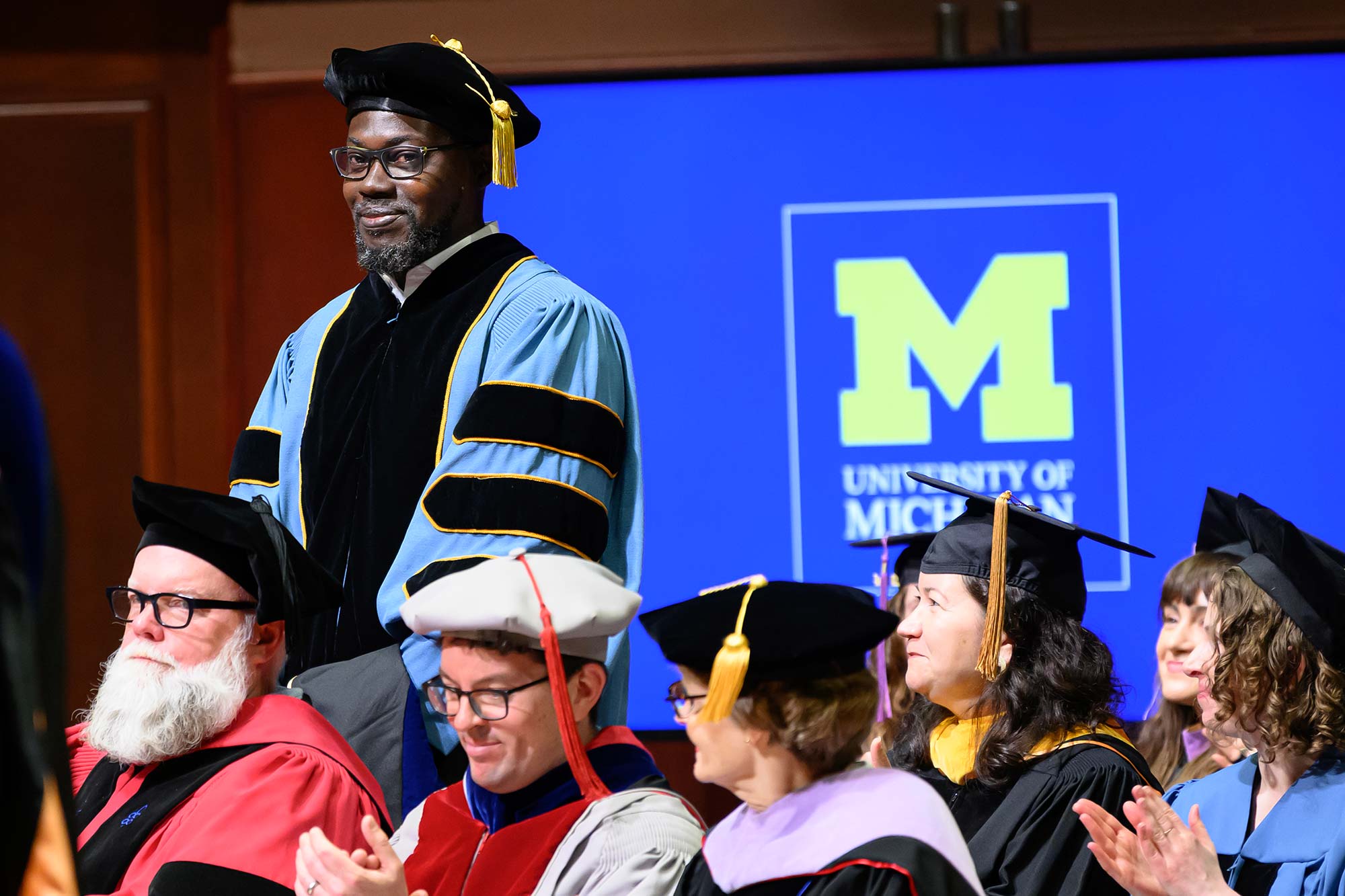 A graduate of Taubman College standing in full regalia in front of a screen with the university logo.