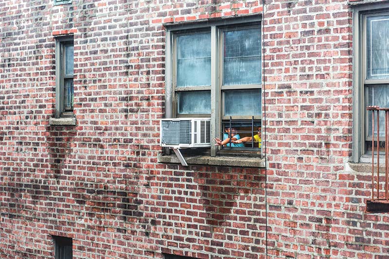 An air conditioner in the window of a brick building with children reaching out an adjoining window.