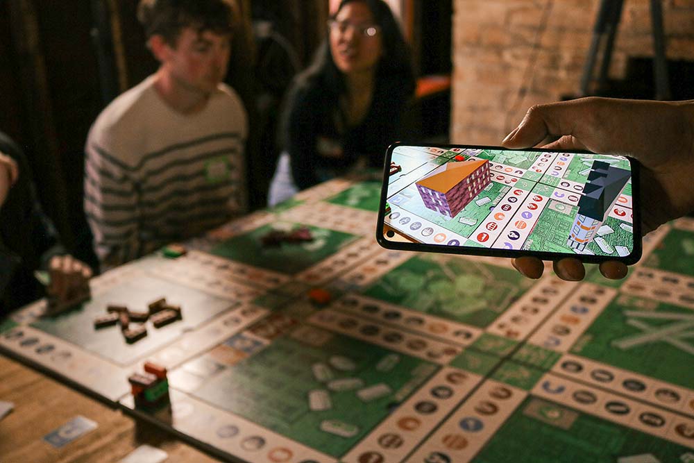 A phone showing an augmented reality interface with virtual buildings overlaid on the game board for Holding Pattern.