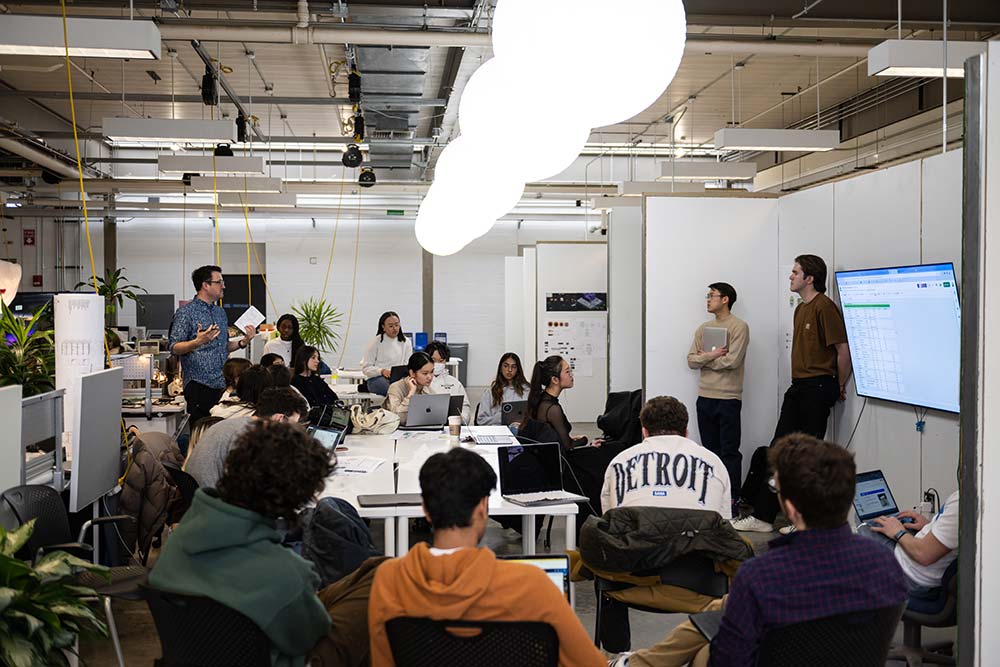 Students sitting around a table listening to two individuals presenting and discussing an image on the television screen.