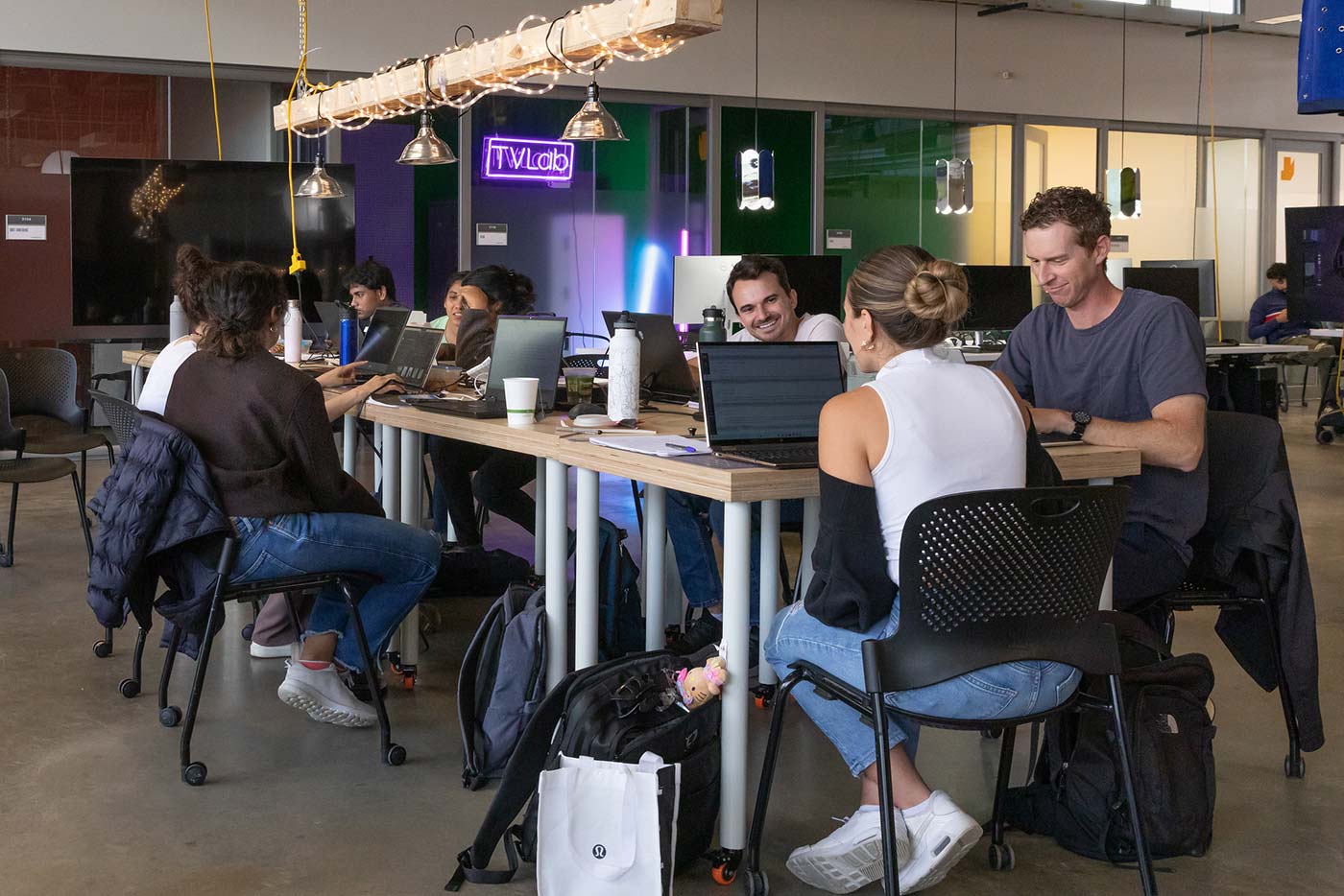 A group of people sitting around a table in Studio Reassembled conversing and working on laptops.