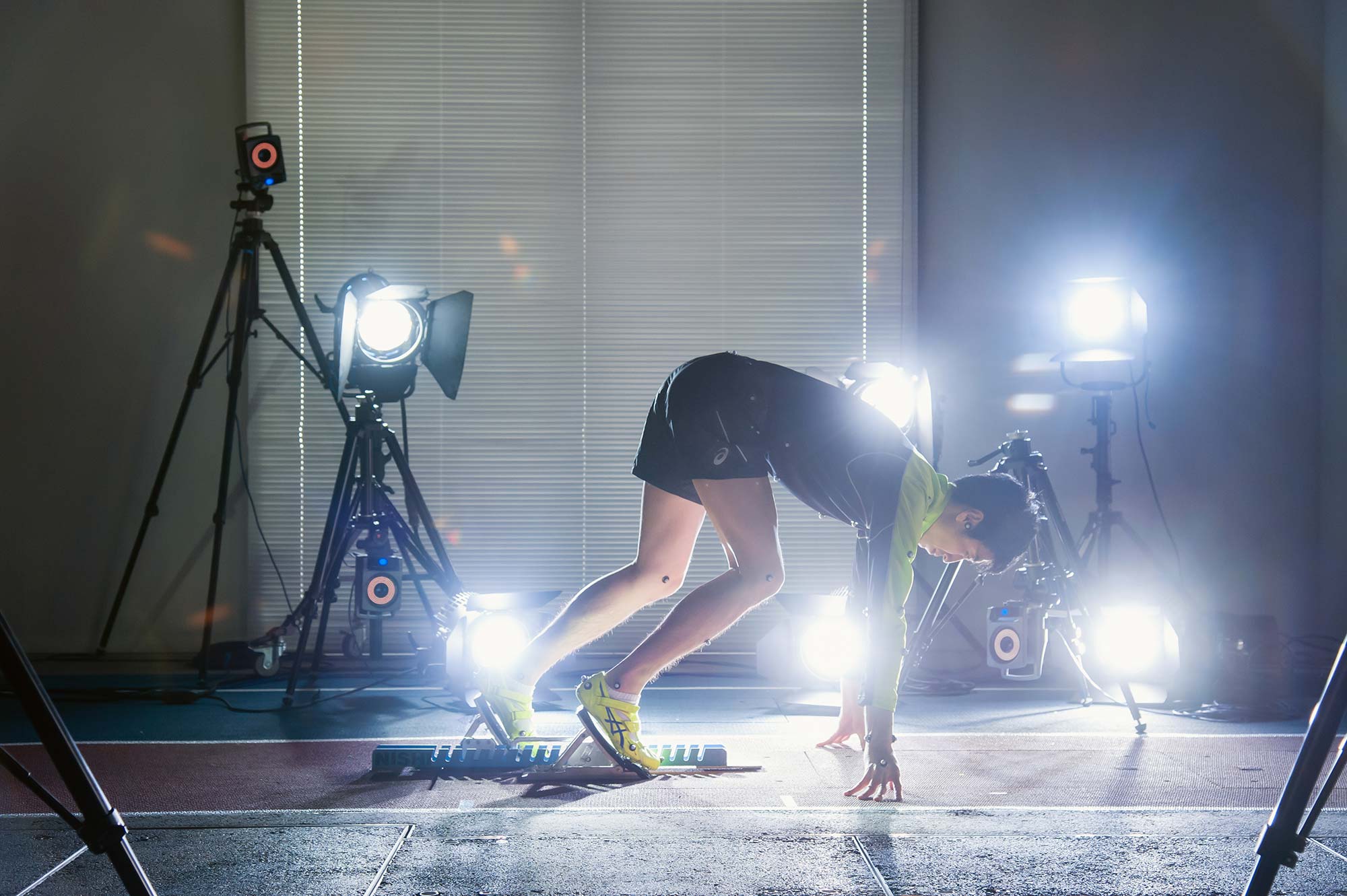 An athlete in running shoes and athletic clothing leans forward in a sprint start position on an indoor track. Bright lights, cameras on tripods, and sensors around the space suggest a controlled sports science or performance testing environment.