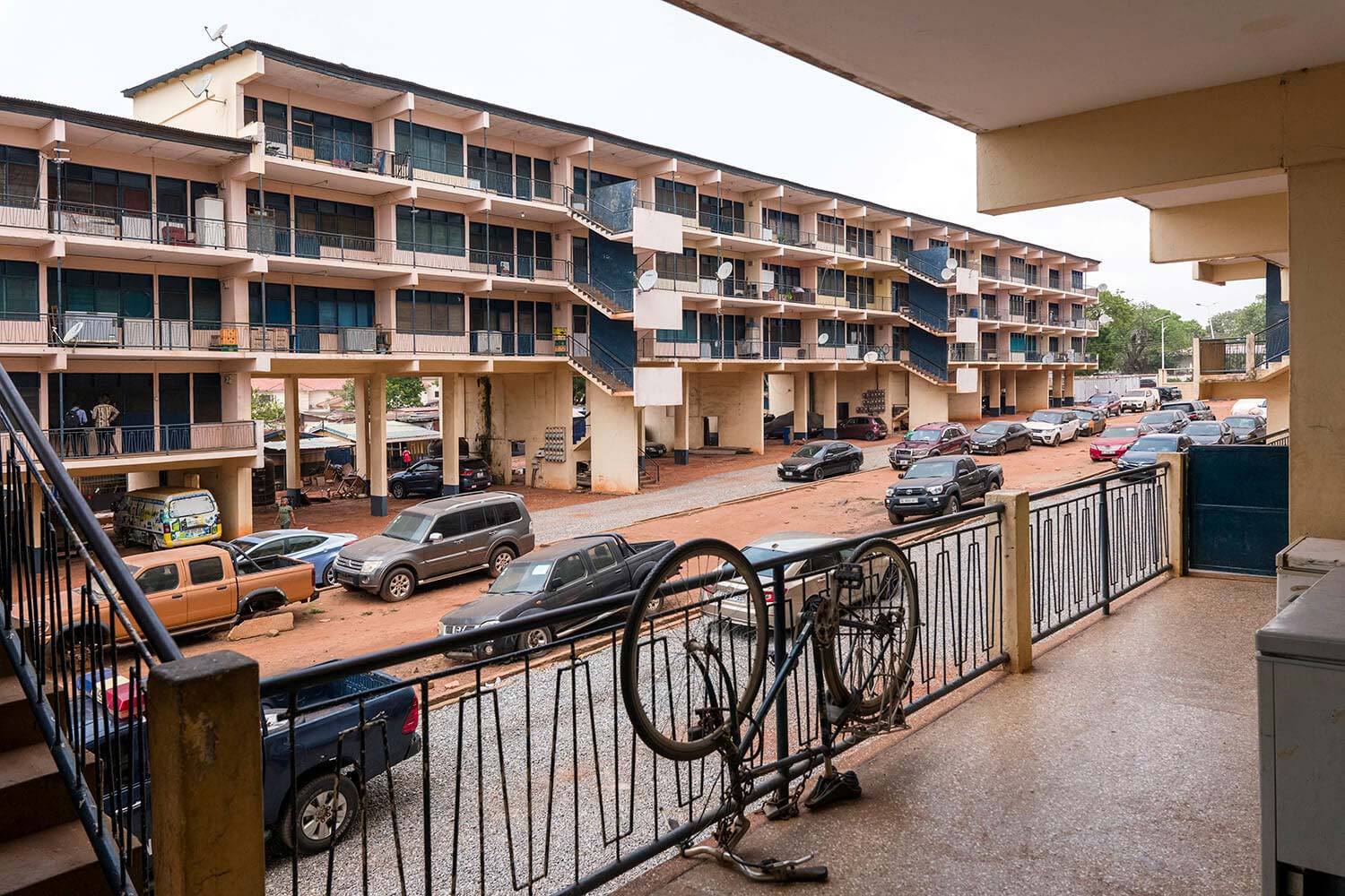 A multi-story apartment building with exterior staircases overlooks a street filled with cars, seen from a balcony with a bicycle leaning against the railing.