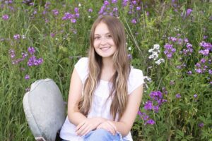 A portrait of Aysha Vandenbosch sitting on a chair in a field of flowers