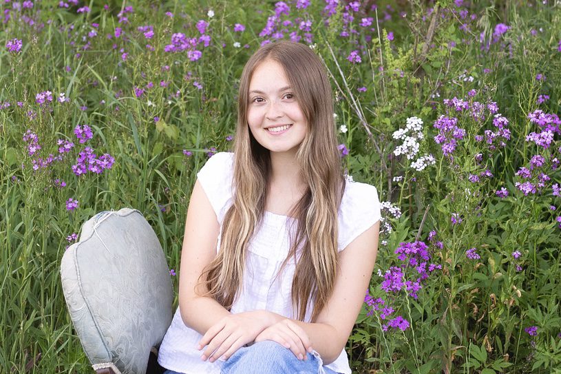 A portrait of Aysha Vandenbosch sitting on a chair in a field of flowers