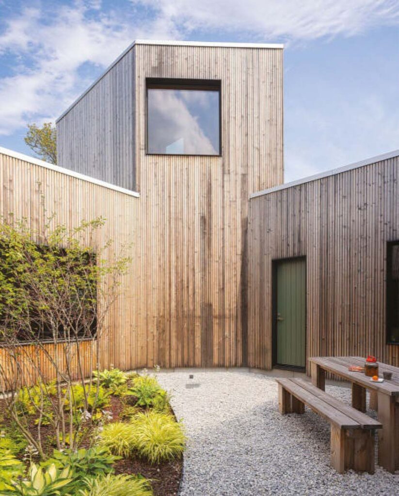A wood clad building courtyard with greenery and a bench and table on gravel.