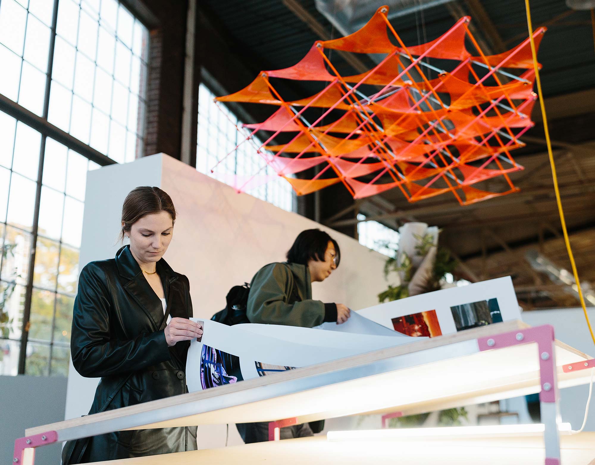 Two people examine large printed images laid out on a table inside a spacious gallery or studio.