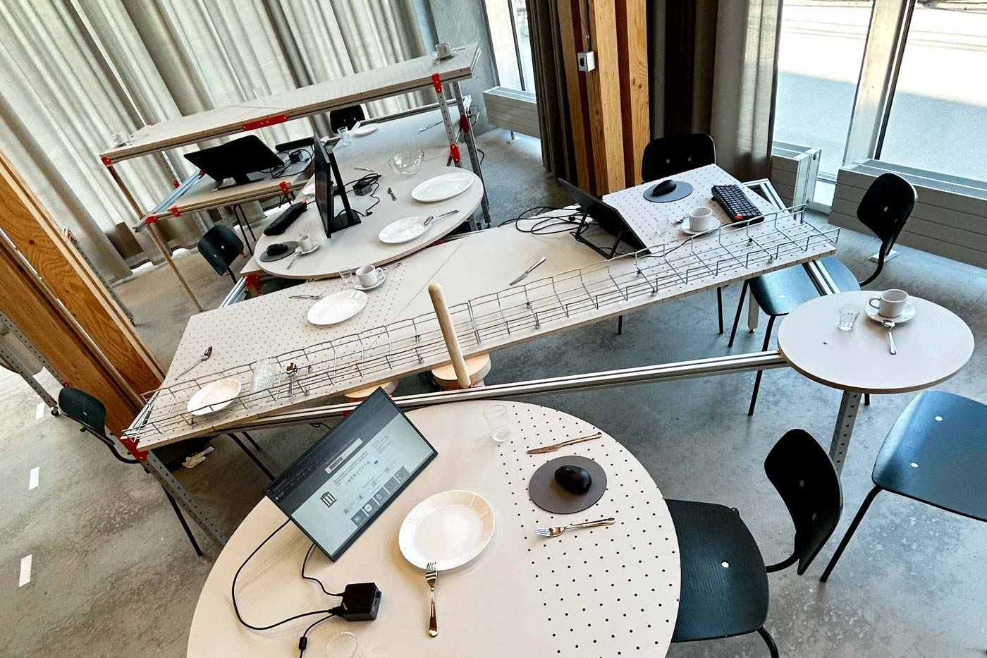 A room with several tables arranged in a zigzag layout, each set with plates, cups, utensils, and computer screens.