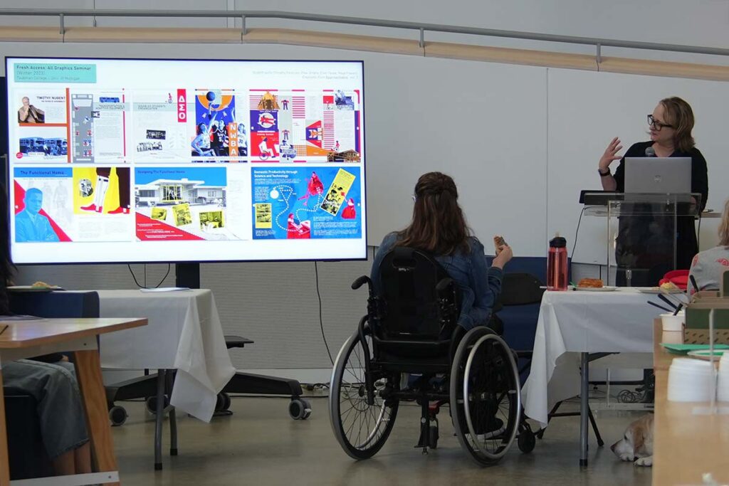 McMorrough speaks beside a screen of advertisement slides as an attendee in a wheelchair faces the front.
