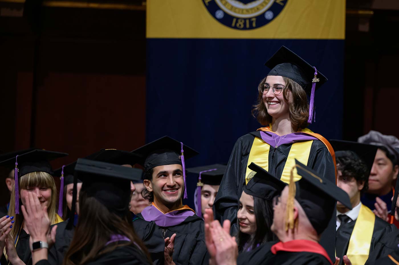 Graduates in caps and gowns applaud as one graduate stands beneath the university banner.