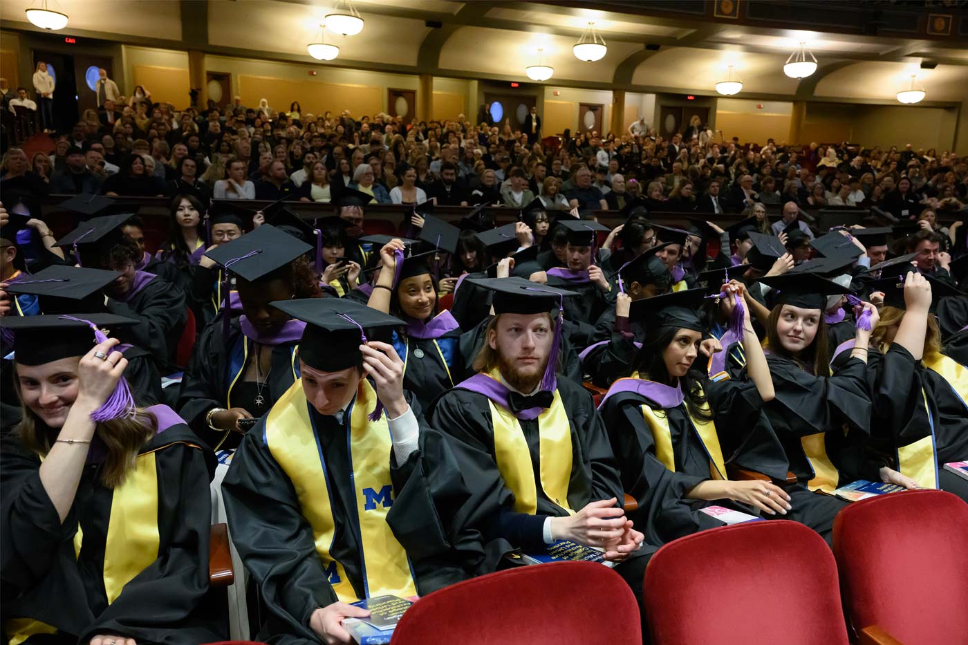 Taubman College graduates in caps and gowns adjust tassels in a crowded auditorium during a ceremony.