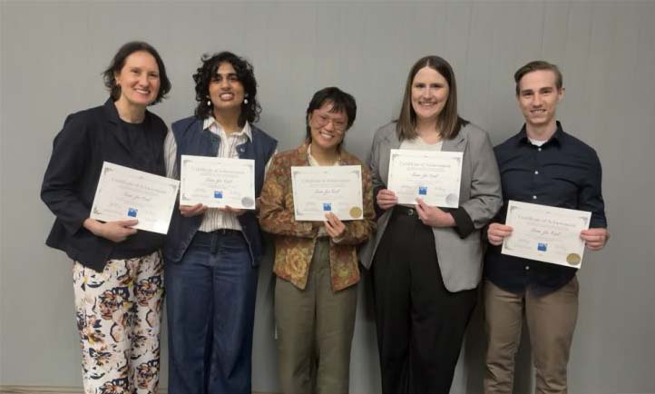 Five students stand side by side holding certificates in front of a plain gray wall.