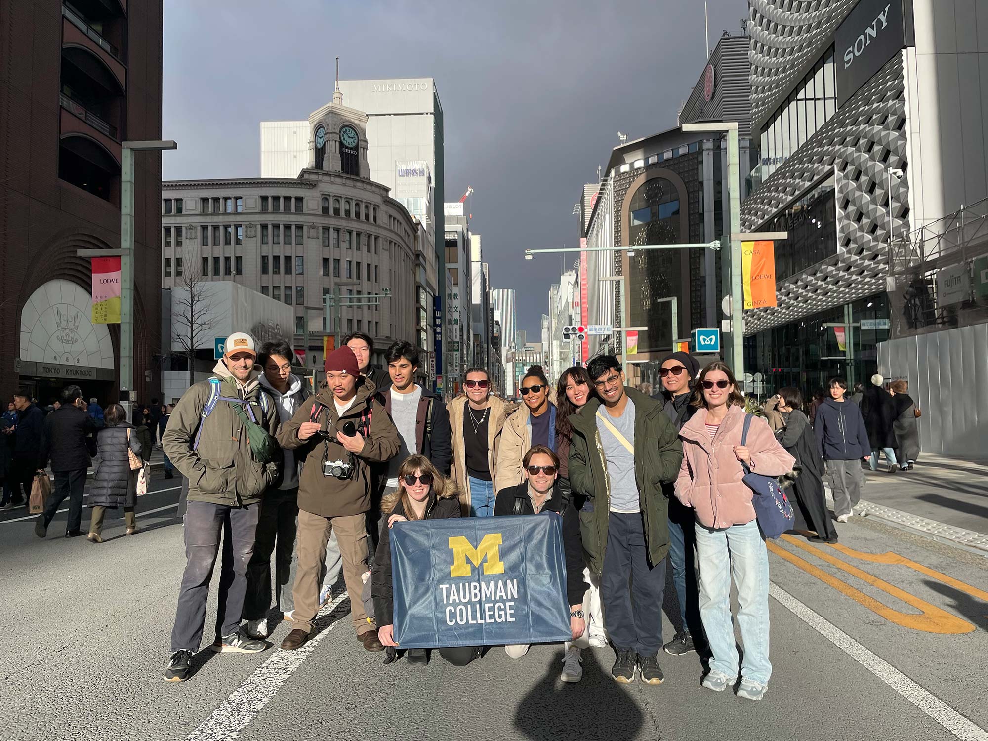 Group poses on a city street holding a “Taubman College” banner with a block M logo.