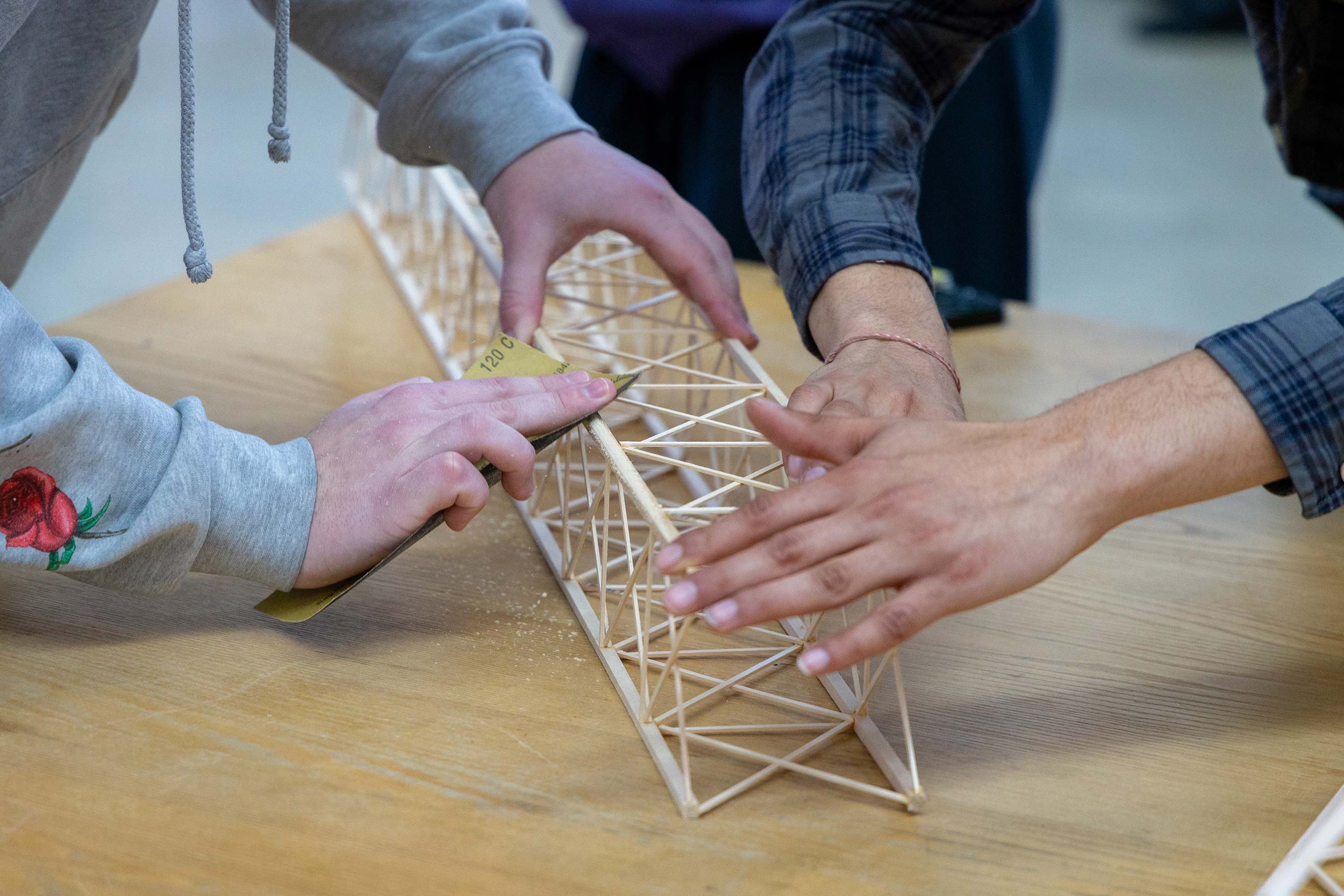 Hands sand and steady a small wooden truss bridge model on a worktable.