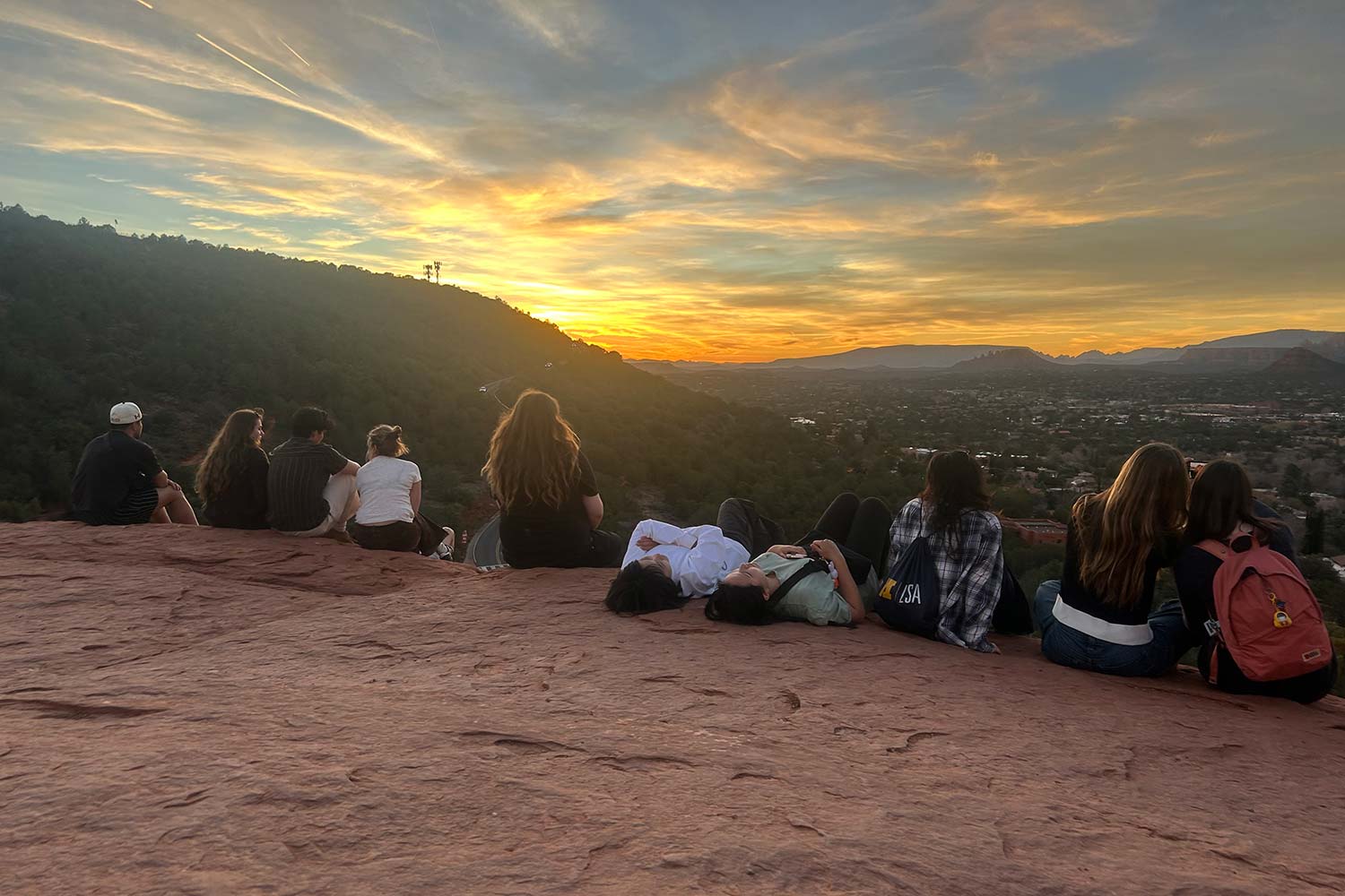 Group of people sit on a red rock overlook watching a sunset over a valley and surrounding hills.